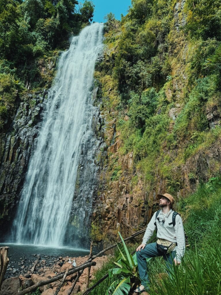 Hiker explores lush waterfall surrounded by dense forest and rocky cliffs.