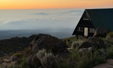 A tranquil sunset scene on Mount Kilimanjaro with a wooden hut overlooking clouds and rocky terrain.