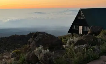 A tranquil sunset scene on Mount Kilimanjaro with a wooden hut overlooking clouds and rocky terrain.