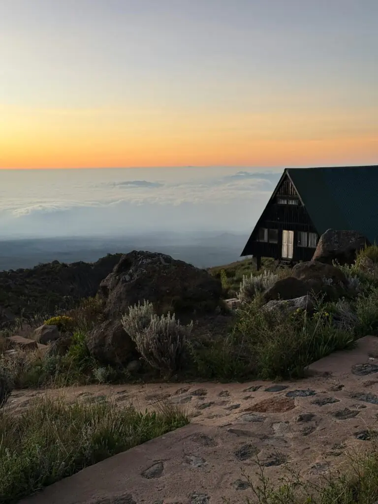 A tranquil sunset scene on Mount Kilimanjaro with a wooden hut overlooking clouds and rocky terrain.