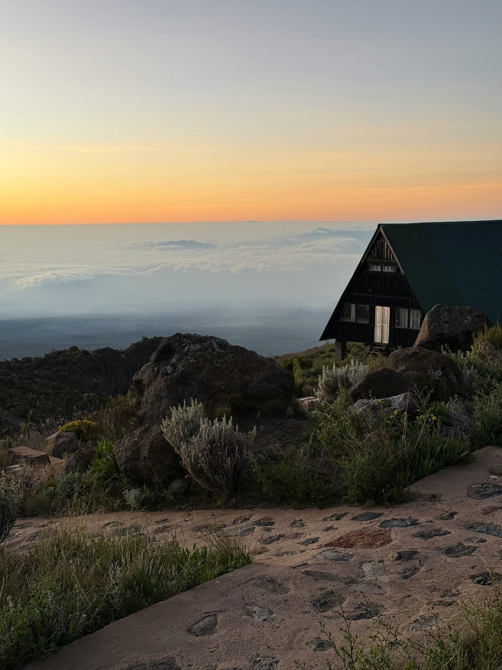 A tranquil sunset scene on Mount Kilimanjaro with a wooden hut overlooking clouds and rocky terrain.