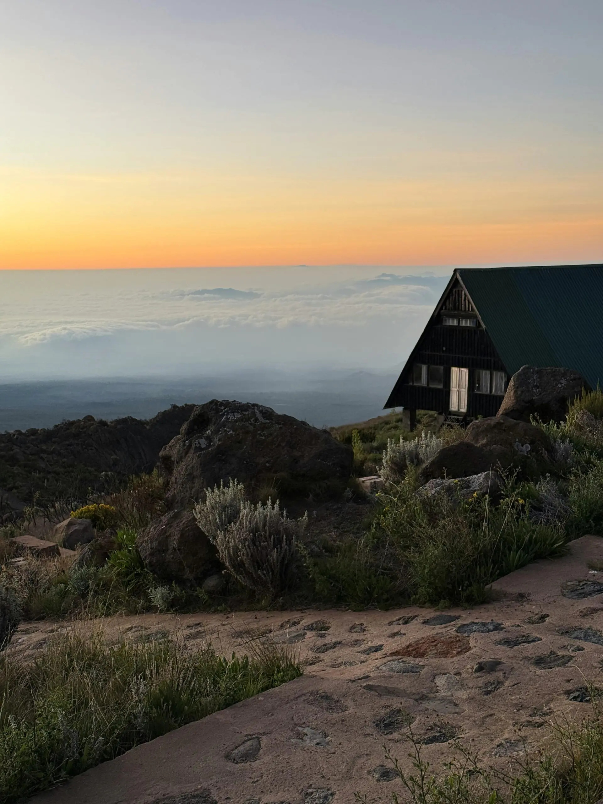 A tranquil sunset scene on Mount Kilimanjaro with a wooden hut overlooking clouds and rocky terrain.
